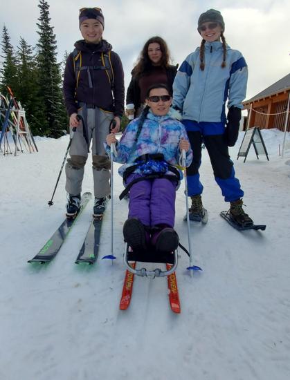 Four individuals pose together on a snowy slope, dressed in winter sports gear. Me, seated in a sitski, wears a colorful jacket and sunglasses. The three standing beside her are holding ski poles and wearing ski & snowshoeing gear. The backdrop features trees and a wooden building.