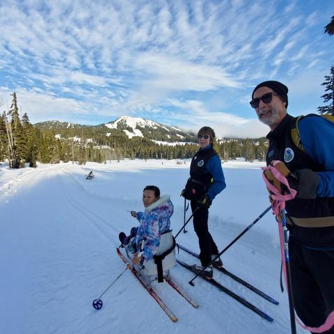 In a snowy landscape, I am sitting in a specialized sitski, smiling with two adults who ski beside her. The mountains and trees create a scenic backdrop under a partly cloudy sky, suggesting a clear, bright day. The individuals are dressed warmly and appear to be enjoying their time together in the winter setting.