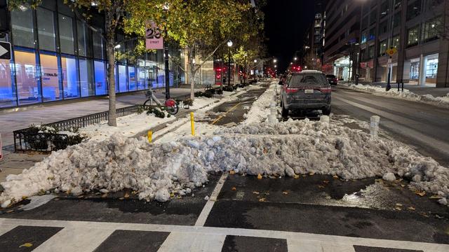 The start of a parking protected bicycle lane a day after a snow storm. The adjacent road and the bike lane are plowed but a 2 foot high bank of snow blocks the entrance to the bike lane.