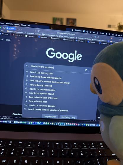 A Pokémon plushie is positioned next to a laptop displaying Google search results. The search bar shows various queries related to self-improvement, including "how to be the very best"