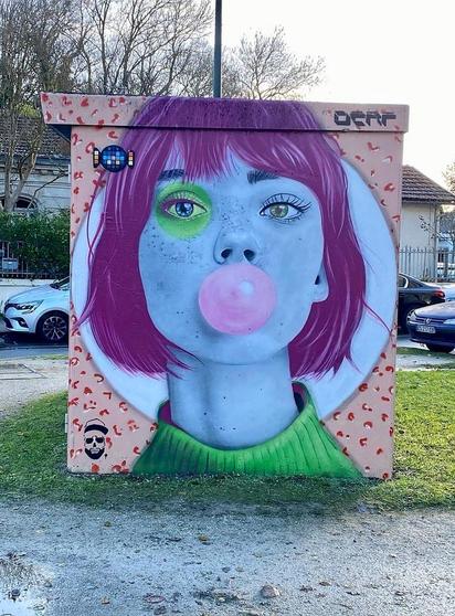 Streetartwall. A cute mural of a girl with chewing gum was sprayed/painted on the outside wall of a small square building (perhaps a bus stop). On the 240x200 wall is the blue face of a young woman with a purple pageboy haircut and green turtleneck sweater, blowing up a pink ball of gum with her mouth. The colorful portrait is framed with a light blue circle and small pink graffiti drawings and one eye with a green circle. In the top corner there is a small candy mosaic made up of different colored rectangles by French artist EssylaR.
