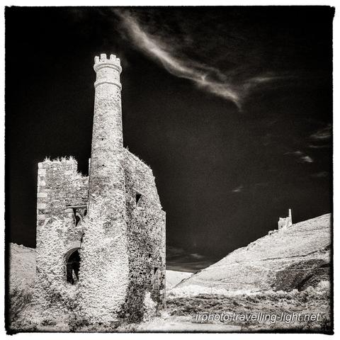 A toned black and white infrared photo of a derelict pumping engine house of a Cornish tin mine with creeper partly covering the walls. A similar building is seen on a hillside in the background.
