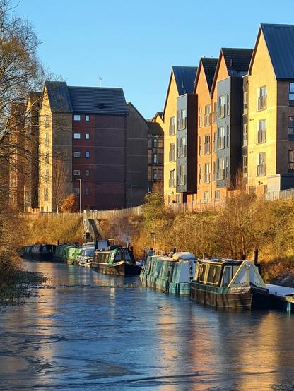 Tall, narrow, gabled modern buildings rising above a canal where a row of boats are berthed.