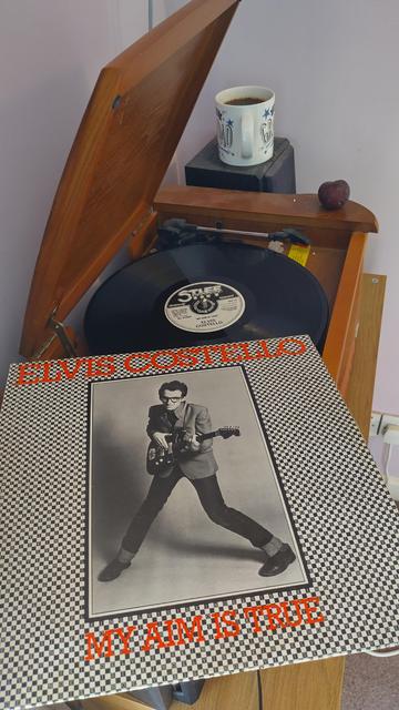 Record player with a 12" black vinyl record
In the foreground is an album cover with a black and white photo of Elvis Costello in a wide legged stance, holding his electric guitar.
Behind the player is a mug of coffee on a speaker. On the corner of the player is a delicious purple plum.