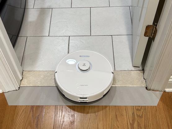 A white RoboRock vacuum robot is entering the laundry room from the hall.  Its back wheels have just cleared the 3D printed ramp that help it climb over the threshold.