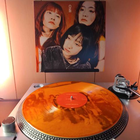 A transparent orange vinyl record sits on a turntable. Behind the turntable, a vinyl album outer sleeve is displayed. The front cover shows the 3 members of Chilli Beans. crowded together and looking in different directions away from the camera.