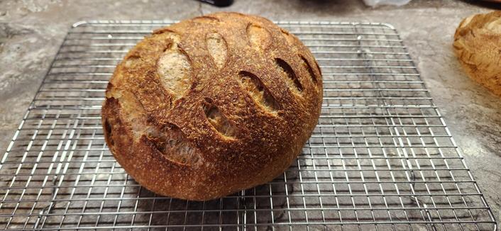A golden brown boule of sourdough bread sitting on a wire rack on a grey kitchen counter.