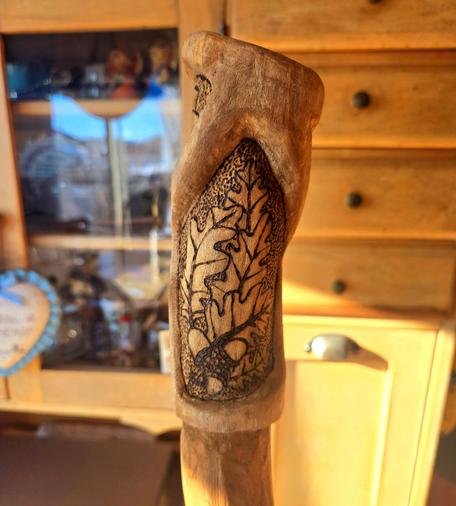 The top of a walking stick that is carved with curved designs. There is wood burned designs of oak leaves