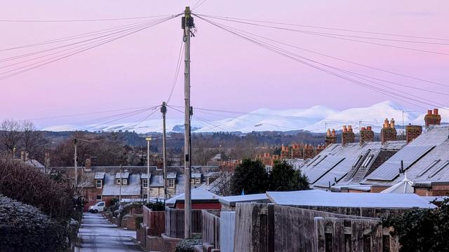 Looking downhill along a lane behind a street of frost covered single story terraced houses towards a range of white snow covered hills in the distance. There are several telephone poles along the lane with black wires leading to the houses. The sky has a slight gradient from peach through pink to lilac where it meets the hills.