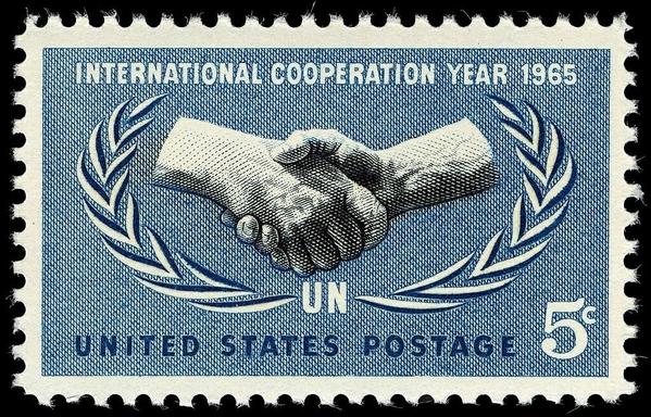 A postage stamp from 1965 features two hands shaking inside a laurel wreath, symbolizing international cooperation under the UN. Bureau of Engraving and Printing. Designed by Herbert M. Sanborn and Olav S. Mathiesen., Public domain, via Wikimedia Commons