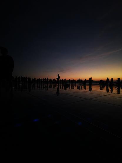 A dark silhouette of a crowd of people stands on a reflective surface at sunset. A single person appears to be playing a saxophone in the crowd. The sky is a mix of dark blue and orange hues from the setting sun. The reflective surface shows the silhouettes of the people and the sky.