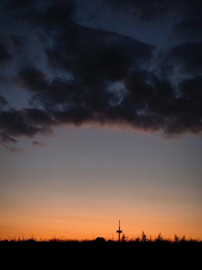 A sunset with a mostly dark sky, featuring a thick band of dark clouds across the middle. Below the clouds, the sky is a gradient of orange and light blue. In the bottom center of the image, a tall, thin silhouette of a structure, possibly a communications tower, is visible against the sunset. Silhouetted grasses and low vegetation are visible at the bottom of the image.