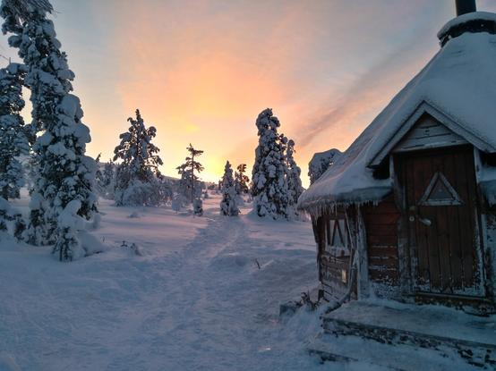 A snow-covered wooden cabin sits in a snowy landscape at sunset. The cabin is dark brown wood with a steeply pitched roof heavily laden with snow and icicles hanging from the eaves. A path leads from the foreground towards the cabin and through a grove of snow-laden evergreen trees. The sky is a blend of orange and pink hues from the setting sun.