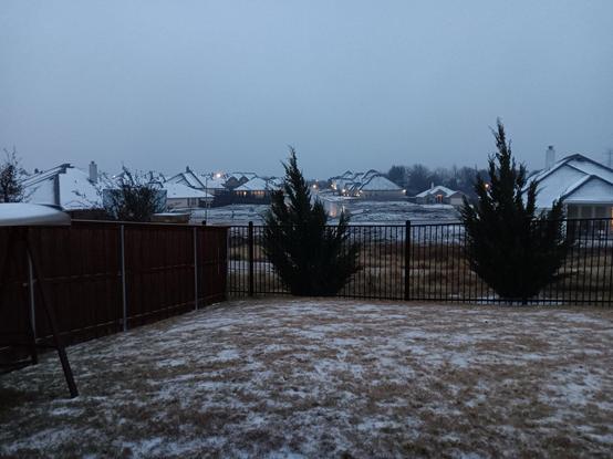 A picture taken from my back porch at dusk of my neighborhood. There is snow mixed in with my grass and the rooftops are also covered with a thin layer of snow.