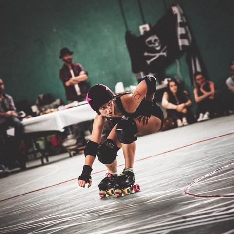 A matte subdued colour picture of a rollerderby playing person skating down low in a corner of the track. In the background spectators are sitting in the floor, a skull and bones flag is visible with a brittany flag beside it.