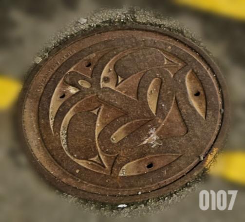 Dark brown maintenance hole cover surrounded by coarse pavement and two partial segments of yellow line paint. The maintenance hole cover features an abstract indigenous design, symmetrical, with many arcs and curved lines. 
The number 0107 appears in the bottom right corner.
