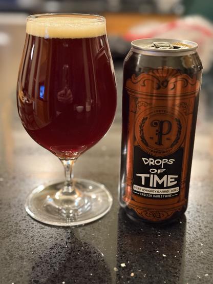 A full tall snifter glass of deep amber colored beer. The pint can is black and copper with white lettering. A script capital “P” is front and center
