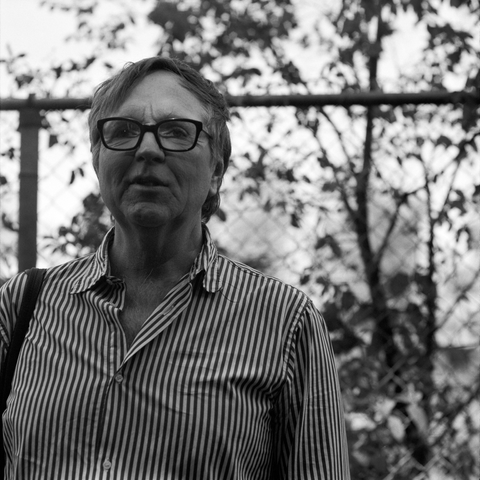 A black and white close up photo of a man standing in dappled shadow before a high chainlink fence. He is looking ahead, past the camera.