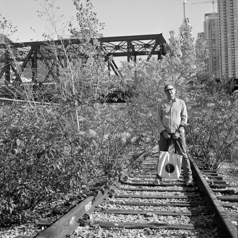 The same man, holding a canvas bag on which is depicted a tomato, stands on a tie of an old railway line, now overgrown with sumac. Behind is a iron bridge and further on some condo towers and part of the CN Tower can be seen.