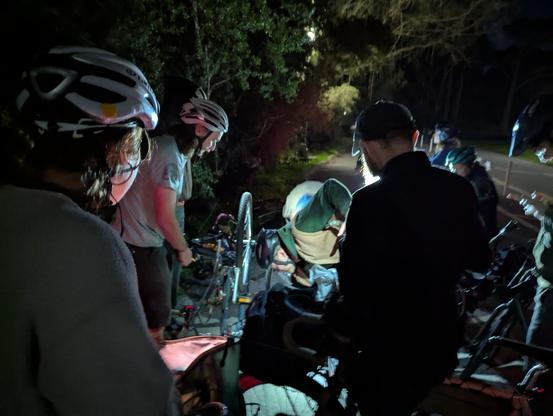 A lathe number of people standing around a bike being repaired in the park at night