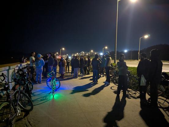 A bunch of people standing around with bikes next to Ocean Beach. One bike has a bunch of blue and green LEDs on it