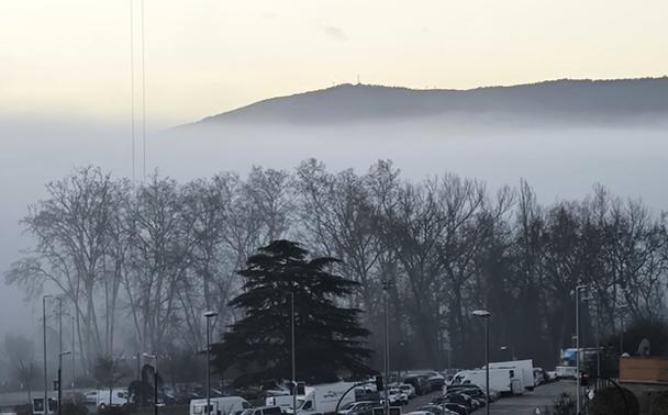 Paisaje en el que al fondo está una sierra, la del Perdón al sur de Pamplona. En primer plano una cadena de árboles sobre un aparcamiento. Entre los árboles y la montaña la niebla