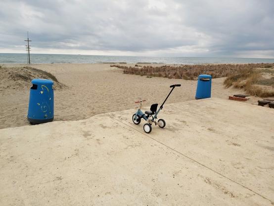 Triciclo infantil en el medio de la imagen, sin niñe, al borde de la entrada de una playa en otoño, con el cielo nublado.