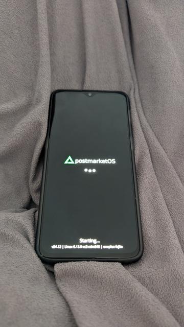 A OnePlus 6T smartphone on a blanket. The display is showing the boot screen with the text "postmarket os" with its logo and the progress dots.
