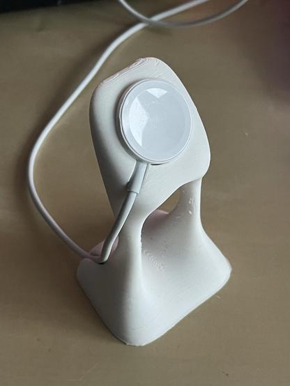 A white accessory stand with a circular charging pad and a cable attached, positioned on a flat surface.