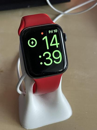 An Apple Watch with a red band displaying the time as 14:39 and the date as Friday 10, resting on a white charging stand. A battery icon indicates charging.