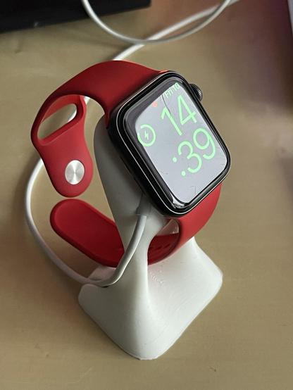 A smartwatch with a black face and bright green display time of 14:39, mounted on a white charging stand. The watch has a red strap.