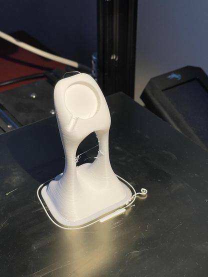 A partially finished 3D print of a small object, featuring a bulbous shape with a narrow neck and a circular top. The print is white and sits on a metal surface, showing strands of filament from the printing process.