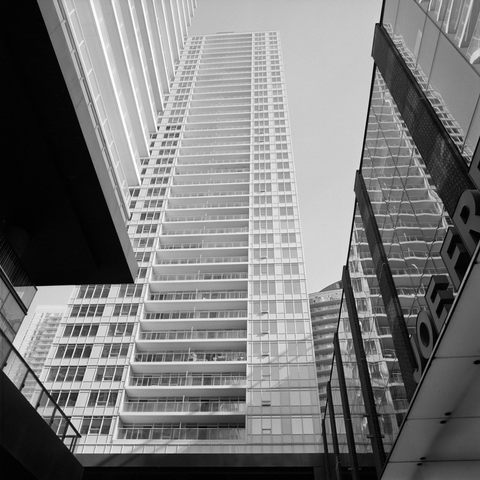 Looking up at a modern white condominium tower from what feels like a deep courtyard surrounded by glass buildings. All but two or three of the many balconies appear barren.