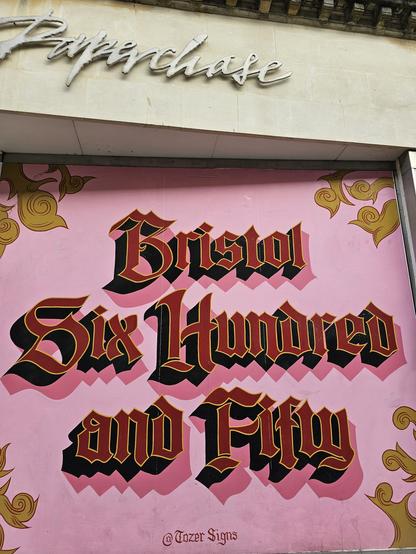 Photograph of a shopfront  overed with board painted pink with red blackletter lettering outlined in gold "Bristol Six Hundred and Fifty". Signed @tozer signs