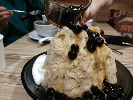 Shaved ice as served in Taiwan: a many hands sized heap of thin flakey shaved ice, tea flavoured, with a cup of glibberish tapioca pearls being poured over it.
