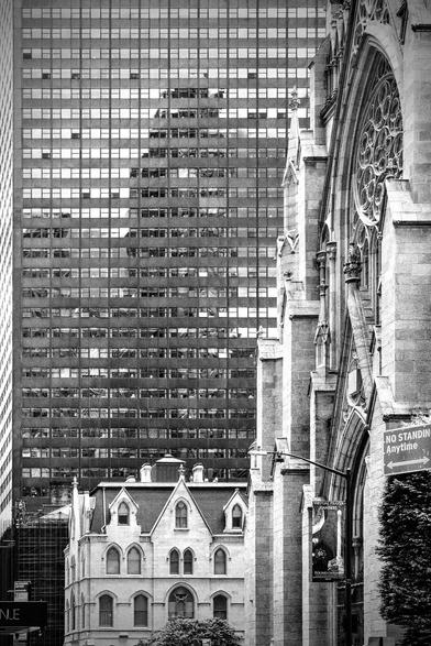 This striking black-and-white photograph captures a juxtaposition of old and new in the heart of Manhattan, New York City. A towering, modern skyscraper, with its grid of reflective windows, dominates the left side of the frame, while on the right, a Gothic-style church stands as a testament to the city's rich history. The church's ornate facade, featuring intricate stonework and a large, arched stained-glass window, is partially obscured by the skyscraper, creating a sense of depth and perspective. In the foreground, a smaller, more residential building with a pitched roof and arched windows adds to the visual complexity of the scene. The overall effect is one of contrast and harmony, as the ancient and the contemporary coexist in this bustling urban environment.
