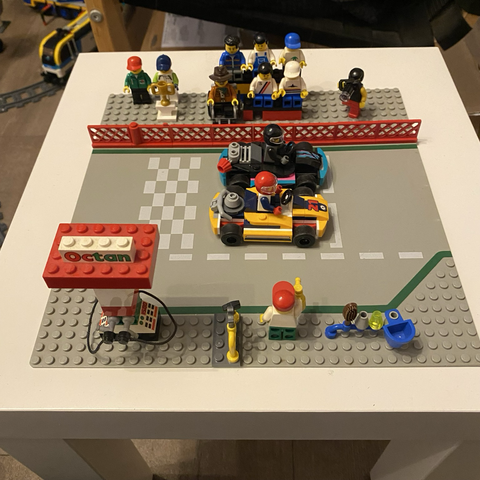Ein Lego-Diorama: Eine Rennstrecken-Basisplatte mit zwei Karts, vorne einer Boxengasse mit Tankstelle und Mechaniker plus Werkzeug; hinter der Strecke eine Tribüne mit Zuschauern, ein Fotograf und einee Rennfahrerin zusammen mit einem Mann in Octan-Shirt vor einem großen Pokal