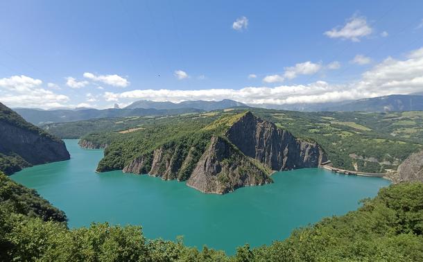 Photographie numérique couleur en format paysage.
Une vue qui surplombe le lac couleur vert entre émeraude et turquoise, avec à droite le sommet du barrage voûte et en face le lac en arc de cercle qui s'étire au loin entre les montagnes abruptes.
Des arbres et des zones vertes aux premier et second plans, une chaîne de montagne bleutée au fond, avec un pic isolé et un ciel bleu bordé de cumulus blancs à l'horizon.