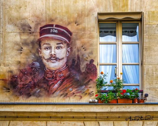 An eye-catching mural adorns the exterior of a Parisian building, depicting a heroic figure in uniform, complete with a detailed mustache. To its right is a window with red potted flowers.