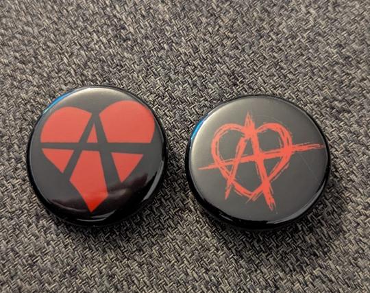 Two black buttons / pins with different styles of the "relationship anarchy" logo on them in red.

The left one depicts the classical, red stylized heart icon with the lines of the anarchy "A" in it in black.

The right one looks basically very close like the original anarchy logo of the "A" and its lines crossing the shape's borders, but instead of a circle, the surrounding shape is a stylized heart-icon drawn in the typical anarchy logo scratched lines.