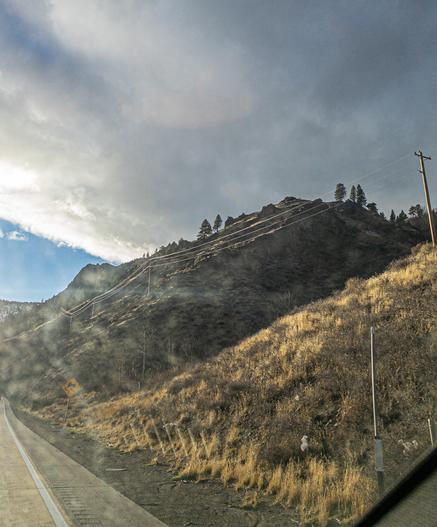From the bottom of the image there is a narrow triangular wedge of the edge of the highway, another triangle of near foothills with power lines running diagonally from left to right. Above is a sky, partly cloudy, with a wedge of clear sky on the far left, and low hanging fair-weather clouds everywhere else.