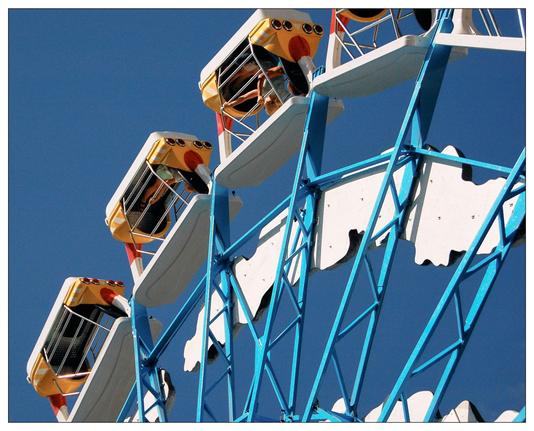 This image features a section of a Ferris wheel set against a clear blue sky. The Ferris wheel has a bold blue and white structure with gondolas that are colored in white with orange and brown detailing. The design elements and color contrast vividly with the blue of the sky, emphasizing the recreational and joyful nature of the ride.