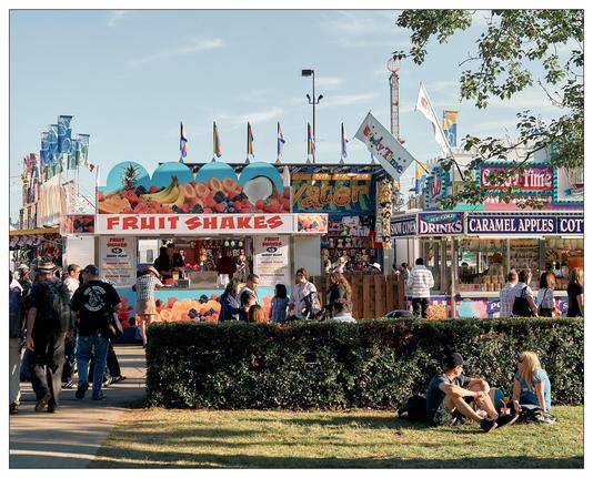 The image shows a bustling fairground with various food and drink stalls. A prominent stall titled "Fruit Shakes" is featured in the foreground, selling fresh fruit shakes, as suggested by images of colorful fruits displayed on its facade. Other stalls visible in the background offer a variety of treats, including "Drinks," "Caramel Apples," "Cotton Candy," and more, highlighting the typical fairground snacks.

The scene is lively, with many people walking around, visiting the stalls, or sitting and relaxing on the grass. There's also a decorative hedge in the foreground, contributing to the festive and orderly environment. In the distance, you can see amusement rides and flags, enhancing the fair's vibrant atmosphere. The setting indicates a sunny day, perfect for outdoor activities like a fair.