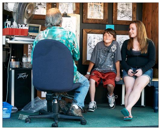 The image shows a scene inside a caricature artist’s booth at a fair or public event. An artist, viewed from behind, is sitting on a rolling chair, wearing a tie-dye top and working on a drawing. Facing the artist are two young individuals, likely waiting for their caricature to be completed. The boy on the left is wearing a dark T-shirt and red shorts, while the girl on the right is dressed in a black sleeveless top and denim shorts. The booth has a casual, cluttered vibe with various items like a fan, a speaker, a cooler, and sketches displayed on the back wall. A paper on the wall indicates the price per person for the drawings. The overall atmosphere is relaxed and cheerful.