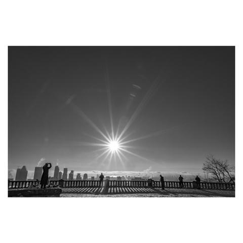 [English follows] Photographie en noir et blanc prise depuis le belvédère Kondiaronk du Mont-Royal. Au centre, un soleil éclatant forme une étoile dans le ciel, ses rayons s'étendant de façon symétrique. Au premier plan, plusieurs personnes en silhouettes sont alignées le long d'une balustrade historique qui projette son ombre sur la neige. En arrière-plan, la skyline de Montréal se découpe avec ses gratte-ciels, et des volutes de fumée s'élèvent dans le ciel froid. À droite, un arbre aux branches nues complète la composition.
Black and white photograph taken from Mount Royal's Kondiaronk Belvedere. In the center, a bright sun creates a perfect star in the sky with symmetrical rays. In the foreground, several people stand in silhouette along a historic balustrade that casts its shadow on the snow. In the background, Montreal's skyline cuts through with its skyscrapers, and wisps of smoke rise in the cold sky. On the right, a bare-branched tree completes the composition.