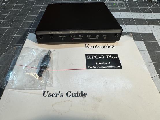 A black electronics box on top of a book that says Kantronics KPC Plus 1200 baud packet communicator User Guide. It is very clean. Not the manual, it is stained. Also there’s a power plug.