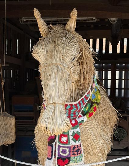 A straw sculpture of a horse wearing a colorful, patterned scarf, set against a rustic background.