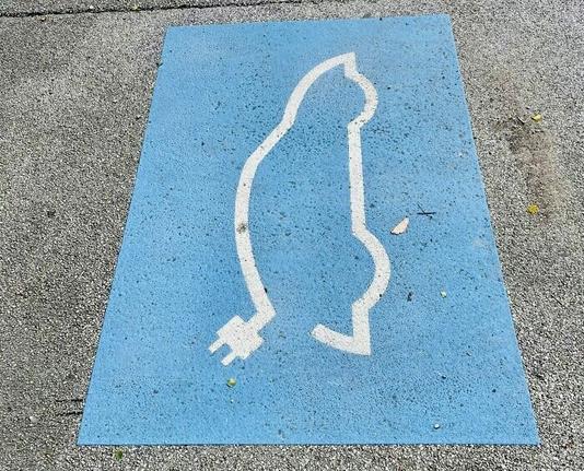 Photo of a road marking that is meant to be the outline of a car with an electric lead coming from it, indicating an EV charging space, however the picture is taken from such an angle that it instead looks like a cat with a charging chord for a tail.