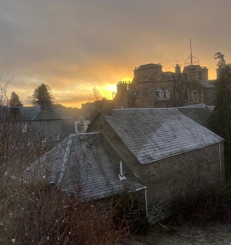 The rear of the Drummond Arms in Crieff with the sun rising over some of the hotel’s chimney pots.  The hotel is in the background with various outbuildings in the foreground.  There are flats in the left of the photo and in the lower left corner bare tree branches with some scattered pink blossom