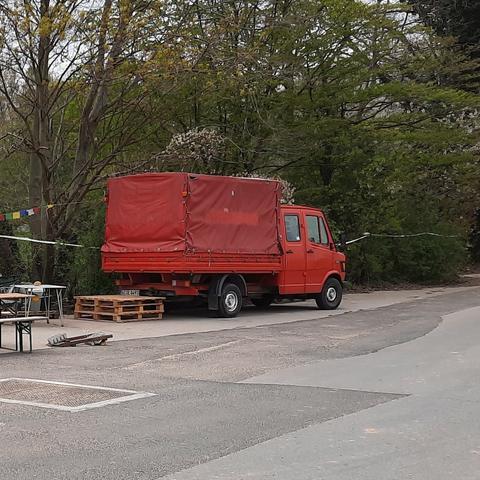 Ein altes, ehemaliges Feuerwehrauto, bekannt als Bündnismobil oder Lauti, steht am Strassenrand neben einigen Bäumen und Büschen. Links von dem Fahrzeug sind einige Paletten und noch weiter links deutet sich mit einigen Tischen und Bänken die Mahnwache an.